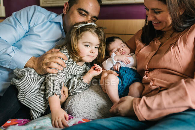 Sonriente padre sosteniendo riendo riendo pequeña hija acostada en la cama con la madre sosteniendo al bebé recién nacido en el fondo en el dormitorio - foto de stock