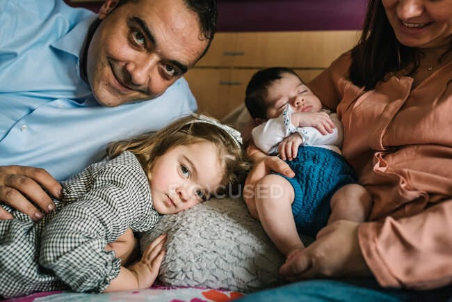 Sorrindo pai segurando rindo pequena filha deitada na cama com a mãe segurando bebê recém-nascido no fundo no quarto — Fotografia de Stock