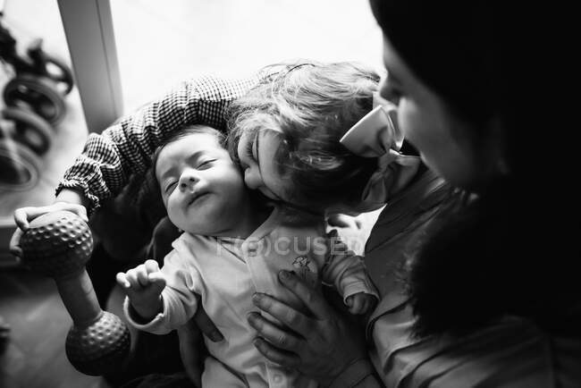 Back view brunette mom hugging cute little baby with her sister kissing at home — Stock Photo