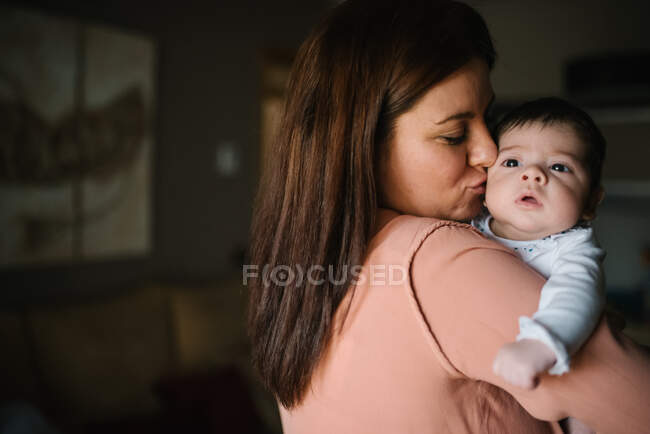 Voltar ver morena mãe abraçando bonito pequeno bebê olhando para longe em casa — Fotografia de Stock