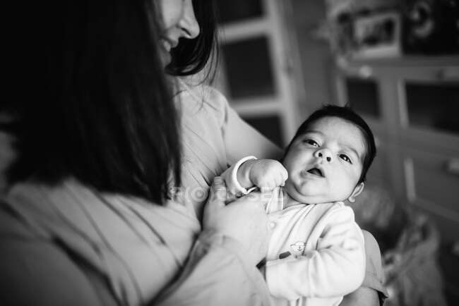 Morena mamá abrazando lindo pequeño bebé mirando lejos en casa - foto de stock