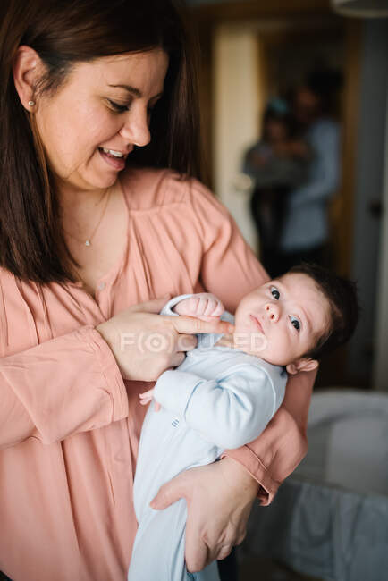 Morena mamá abrazando lindo pequeño bebé mirando lejos en casa - foto de stock