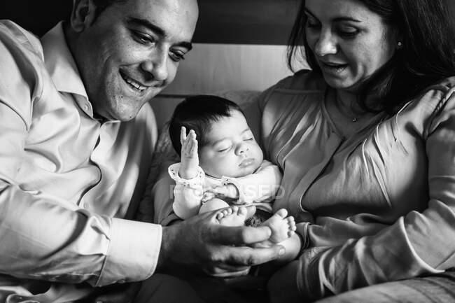 Sonriente padre sosteniendo riéndose hija pequeña acostada en la cama con la madre sosteniendo bebé recién nacido - foto de stock