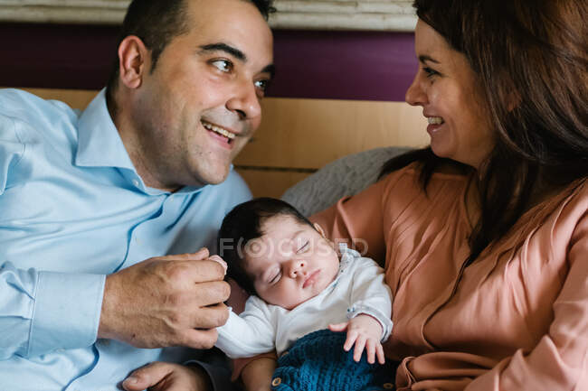 Sonriente padre sosteniendo riéndose hija pequeña acostada en la cama con la madre sosteniendo bebé recién nacido - foto de stock
