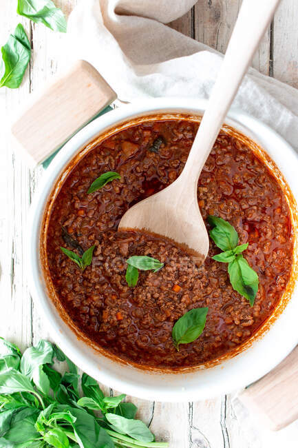 From above white ceramic pan with ragout Bolognese and spoon placed on wooden table with towel and basil — Stock Photo