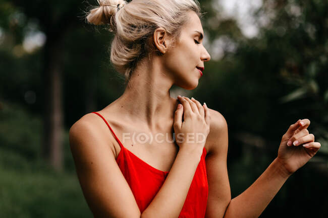 Боковой вид привлекательной блондинки, одетой в красную чувственную позу и трогающей шею с закрытыми глазами среди зеленых цветущих деревьев — стоковое фото