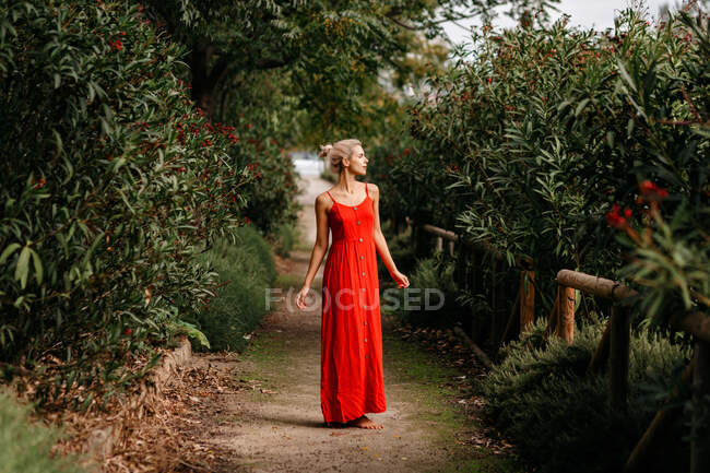 Vista lateral de loira atraente vestida de vermelho sensualmente posando com olhos fechados entre árvores verdes florescendo — Fotografia de Stock