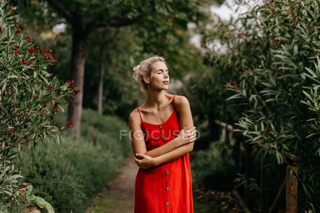 Side view of attractive blonde dressed in red sensually posing with closed eyes among green blooming trees — Stock Photo