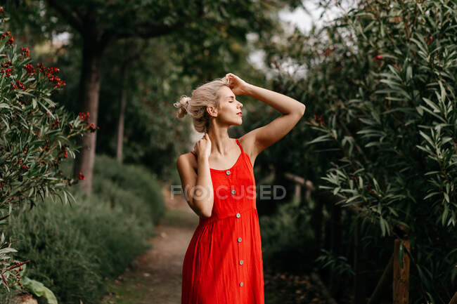 Боковой вид привлекательной блондинки, одетой в красное чувственное позирование с закрытыми глазами среди зеленых цветущих деревьев — стоковое фото