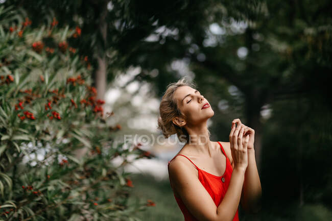 Vista lateral de la atractiva rubia vestida de rojo posando sensualmente con los ojos cerrados entre los árboles florecientes verdes - foto de stock