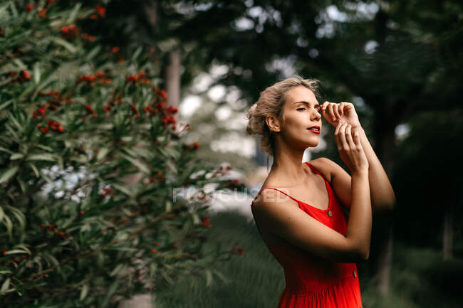 Vue latérale d'une jolie blonde vêtue de rouge posant sensuellement les yeux fermés parmi les arbres à fleurs vertes — Photo de stock