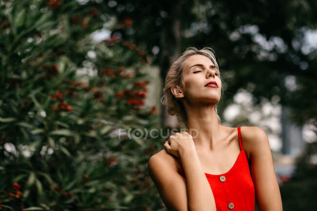 Vista lateral de loira atraente vestida de vermelho sensualmente posando e tocando seu pescoço com olhos fechados entre árvores verdes florescendo — Fotografia de Stock
