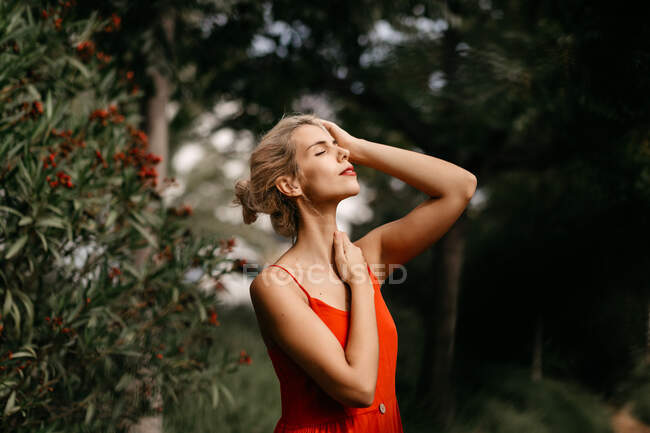 Side view of attractive blonde dressed in red sensually posing and touching her neck with closed eyes among green blooming trees — Stock Photo