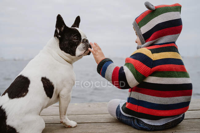 Back view of unrecognizable kid in striped jacket patting spotted French Bulldog while sitting on lumber pier near sea — Stock Photo