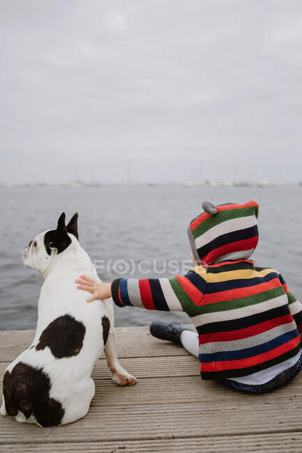 Vue arrière d'un enfant méconnaissable en veste rayée tapotant repéré Bouledogue français assis sur une jetée de bois près de la mer — Photo de stock
