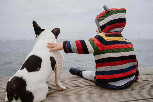 Back view of unrecognizable kid in striped jacket patting spotted French Bulldog while sitting on lumber pier near sea — Stock Photo