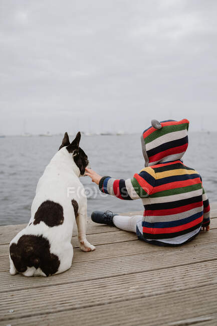 Rückansicht eines nicht wiederzuerkennenden Kindes in gestreifter Jacke, das auf einem Holzsteg am Meer sitzt und die französische Bulldogge tätschelt — Stockfoto