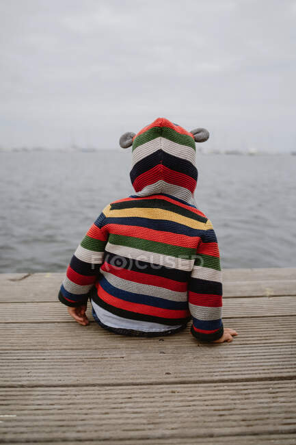 Visão traseira de criança irreconhecível em jaqueta listrada sentado no cais de madeira perto do mar — Fotografia de Stock