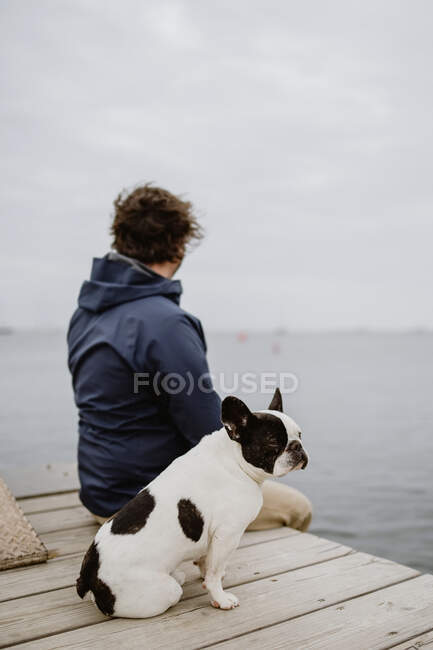 Anonymous adult male in warm jacket embracing spotted French Bulldog while sitting on wooden pier and admiring view of rippling sea on dull day — Stock Photo