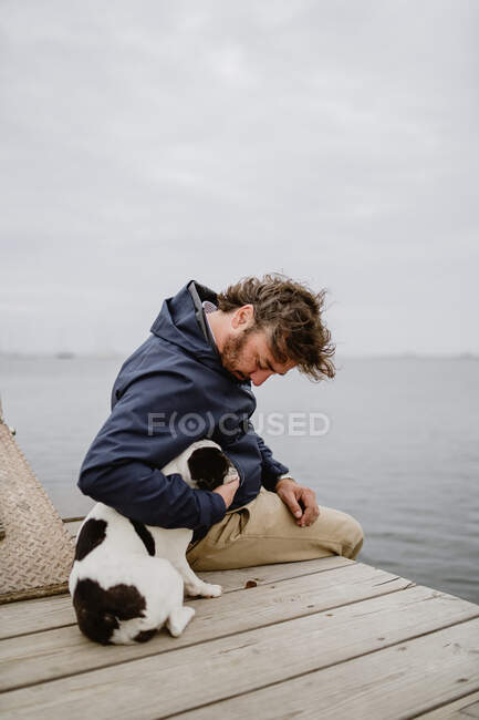 Erwachsene Männchen in warmer Jacke umarmen gefleckte Französische Bulldogge, während sie auf einem hölzernen Steg sitzen und den Blick auf das plätschernde Meer an einem trüben Tag bewundern — Stockfoto