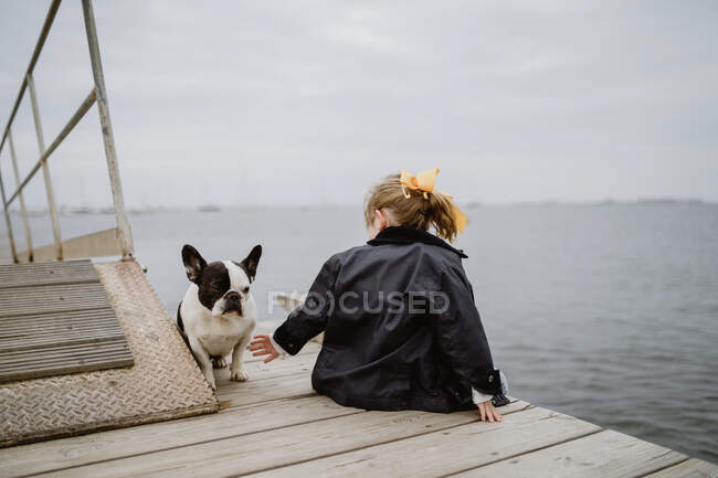 Вид сзади на маленькую девочку с французским бульдогом, сидящую на пирсе у моря в тусклый облачный день — стоковое фото