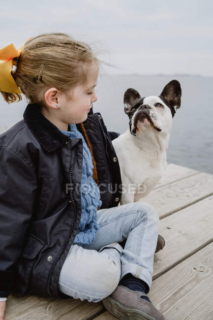 Маленькая девочка с французским бульдогом сидит на пирсе у моря в скучный облачный день — стоковое фото