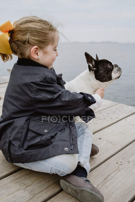 Menina com Bulldog francês sentado no cais perto do mar em dia nublado maçante — Fotografia de Stock
