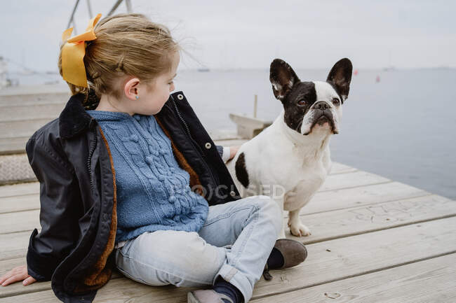 Menina com Bulldog francês sentado no cais perto do mar em dia nublado maçante — Fotografia de Stock