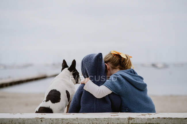Задний вид на двух детей, обнимающих французский бульдог, сидящих на пляже рядом с морем вместе — стоковое фото