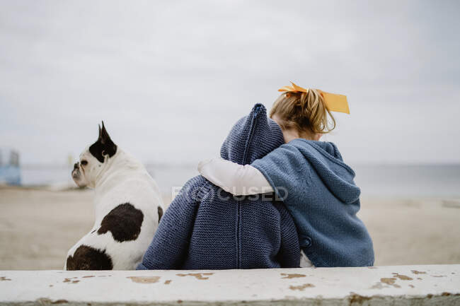 Vista posteriore di due bambini che abbracciano Bulldog francese seduti sulla spiaggia vicino al mare insieme — Foto stock