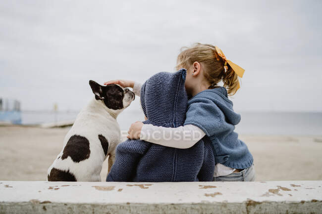 Back view of two children embracing French Bulldog while sitting on beach near sea together — Stock Photo