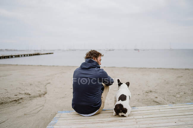 Hombre adulto con chaqueta abrigada abrazando a Bulldog francés manchado mientras está sentado en el muelle de madera y admirando la vista del mar ondulante en un día aburrido - foto de stock