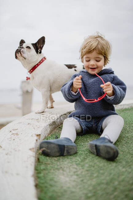 Adorable child smiling and playing with sunglasses while sitting on beach near spotted French Bulldog — Stock Photo