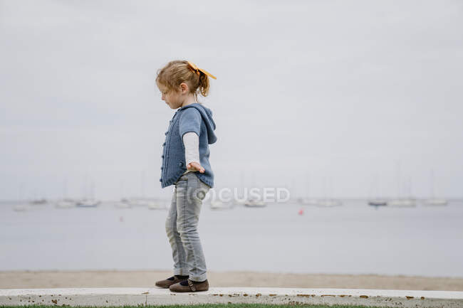 Vue latérale de la petite fille avec les bras tendus marchant sur la frontière contre la mer calme et le ciel gris — Photo de stock