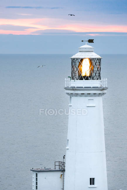 Faro blanco con lámpara de iluminación y flecha en el techo al atardecer en el mar sereno en Gales - foto de stock