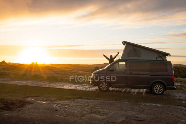 Silhouette féminine avec les mains ouvertes debout sur caravane remorque sur route de campagne solitaire au coucher du soleil au Pays de Galles — Photo de stock