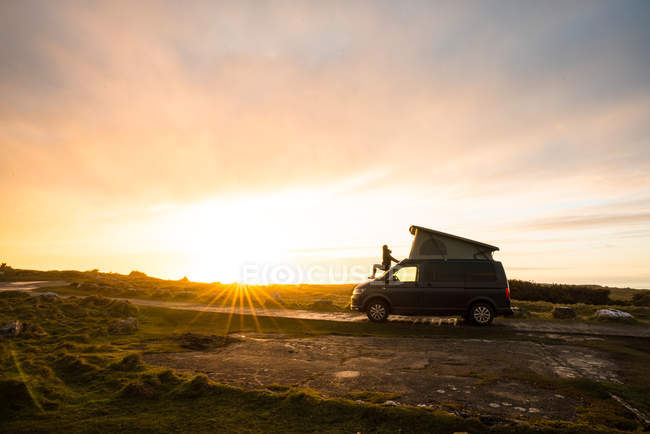 Женский силуэт, сидящий на трейлере каравана на одинокой проселочной дороге на закате в Уэльсе — стоковое фото