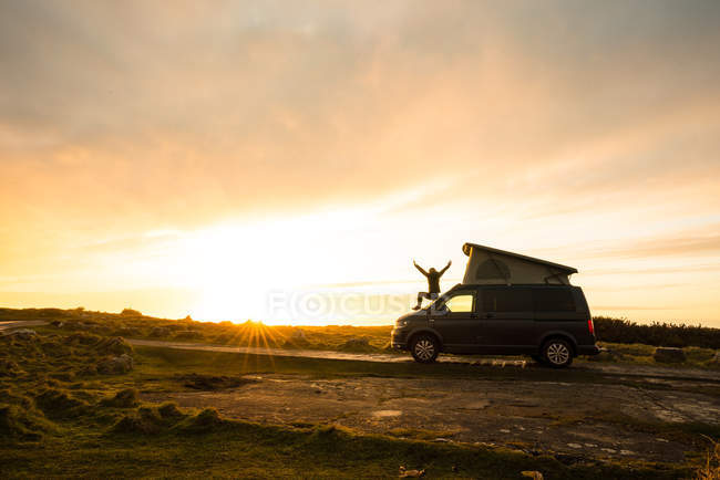 Silhueta feminina com as mãos abertas em pé no trailer da caravana na estrada rural solitária ao pôr do sol no País de Gales — Fotografia de Stock