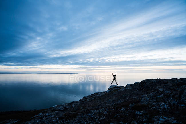 Silhueta de mulher feliz pulando na costa rochosa ao pôr do sol no País de Gales — Fotografia de Stock