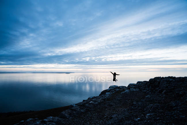 Silhouette de femme heureuse sautant sur la côte rocheuse au coucher du soleil au Pays de Galles — Photo de stock