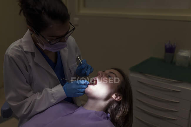 Femme stomatologue en masque et lunettes examinant les dents de la jeune femme avec des outils de dentiste dans la clinique de stomatologie — Photo de stock