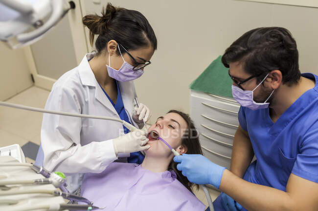 De arriba joven dentista en la máscara y los cristales con el ayudante que trata los dientes de la mujer en la clínica estomatológica - foto de stock
