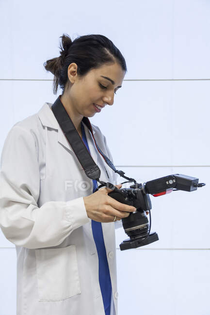 Side view of cheerful young female dentist in medical gown standing in dental cabinet and checking photos on camera — Stock Photo