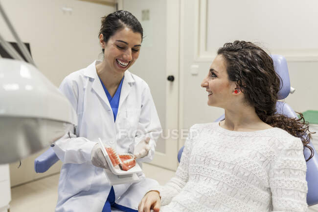 Happy young female stomatologist sitting in dental cabinet. showing dummy jaw to joyful young woman and laughing — Stock Photo