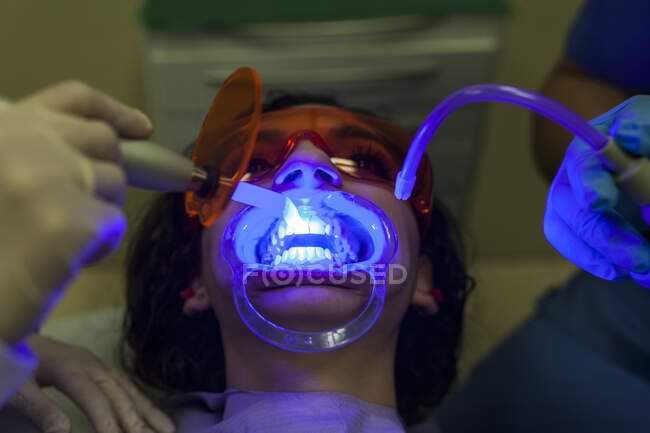 Стоматолог и ассистент осматривают и делают ультразвуковое очищение зубов молодой пациентки со специальным оборудованием с голубым светом в стоматологии — стоковое фото