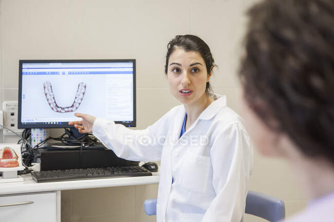 Молода жінка-стоматолог в медичній сукні сидить на стільці і показує зуби пацієнта на комп'ютері в стоматології — стокове фото
