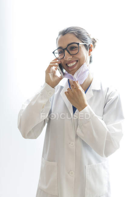 Молода щаслива красива жінка-стоматолог в медичній сукні та окулярах, що розмовляють по телефону і дивиться ізольовано на білому тлі — стокове фото