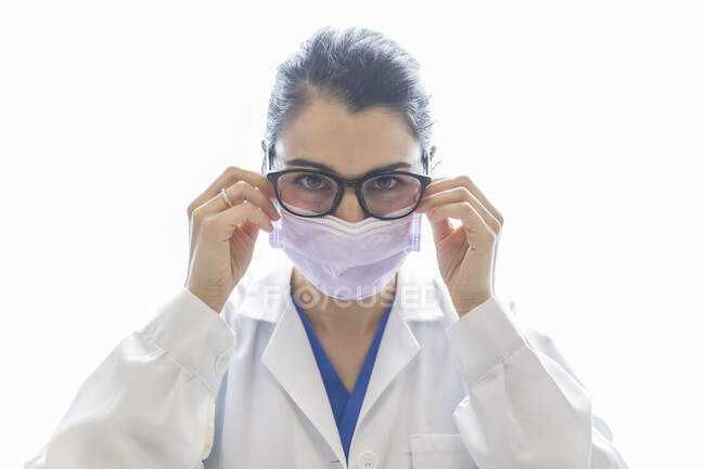 Joven y atractiva estomatóloga con anillo en bata médica, máscara y gafas mirando a la cámara aislada sobre fondo blanco - foto de stock