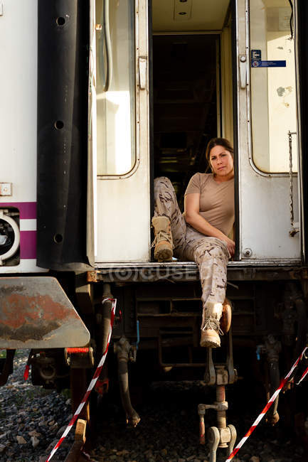 Strong female soldier looking at camera while sitting in doorway of grungy military vehicle in countryside — Stock Photo