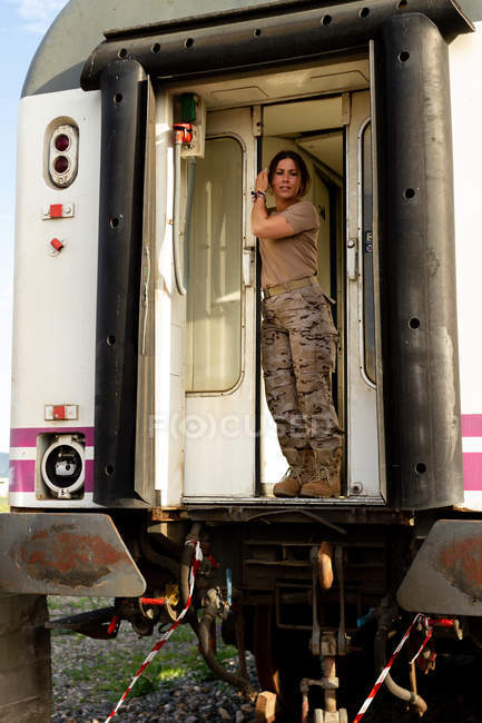 Strong female soldier looking away and smiling while standing in doorway of grungy military vehicle in countryside — Stock Photo
